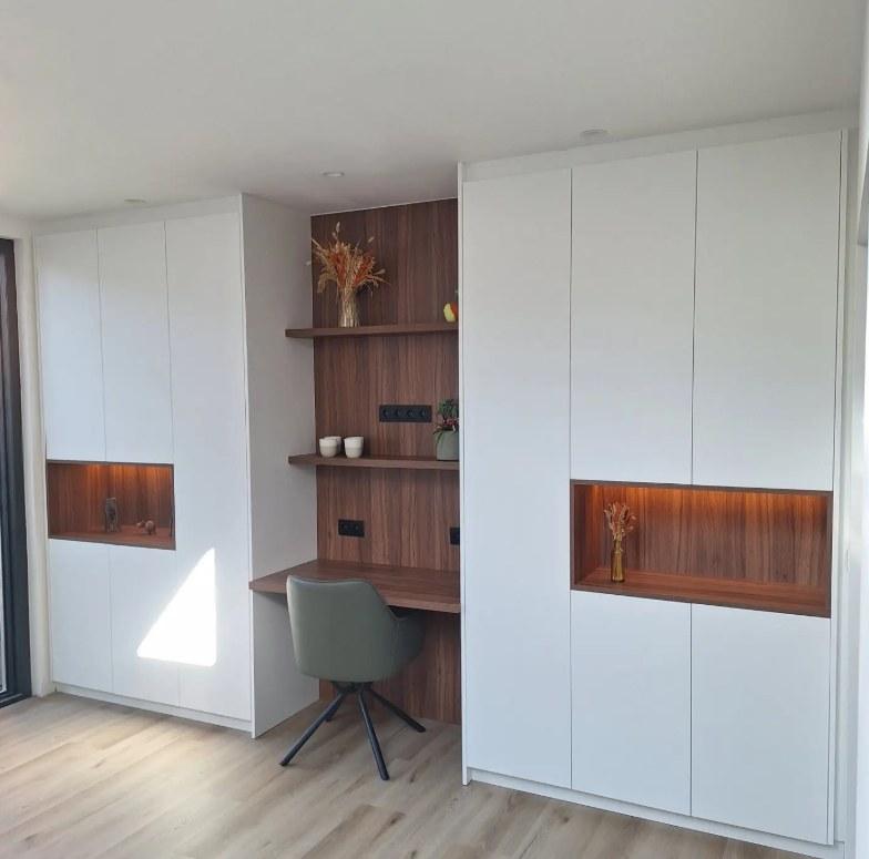 Wand met witte maatkasten, verlichte nissen en een bureau in notelaar